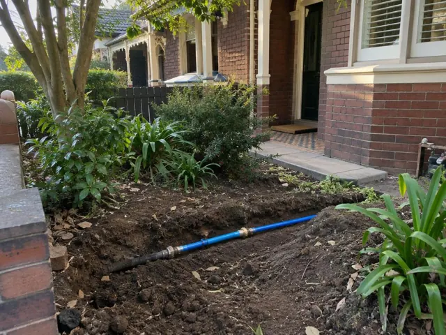 Exposed repaired water pipe in a garden trench beside a brick house in Cammeray