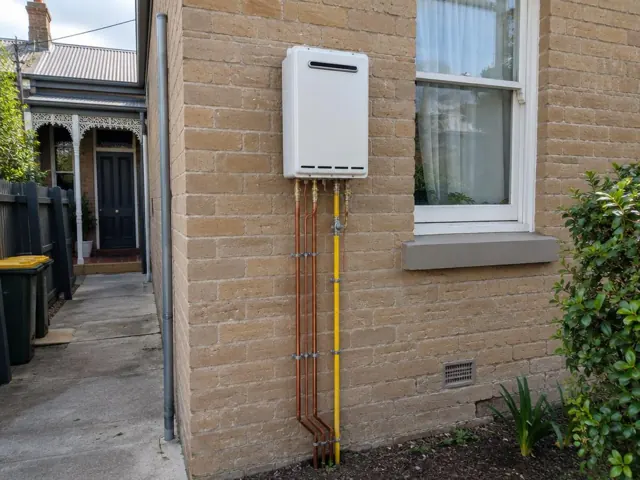 Outdoor wall-mounted hot water unit with copper and yellow pipework on a brick house exterior in Waverton